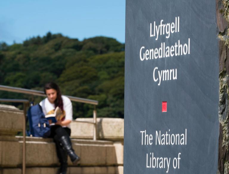 Girl reading on step beside sign for the national library of Wales