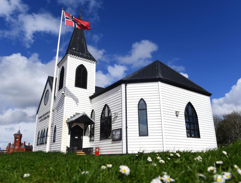 Image of a white Norwegian church and a Norwegian flag with grass in the foreground