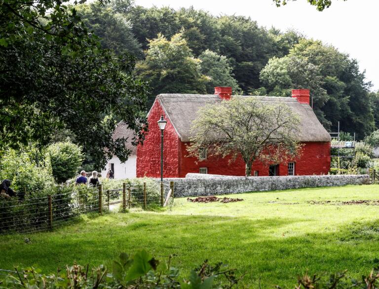 farmhouse building painted red with thatched roof in background with green field in foreground.