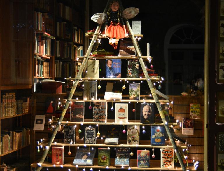 Night time photo of bookshop window, with books on Christmas tree shaped shelving with lights  and toy fairy at the top.