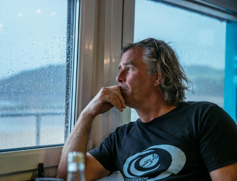 Side profile of surfer Pete Jones looking out of a rain-streaked window at the Gower coastline