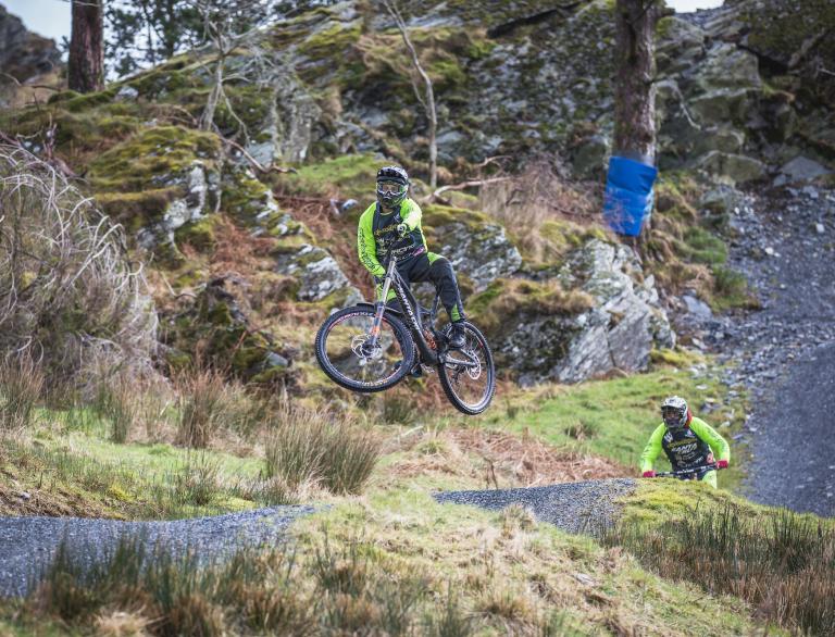 Two bikers on a mountain bike trail, one of which is soaring in the air after a jump.