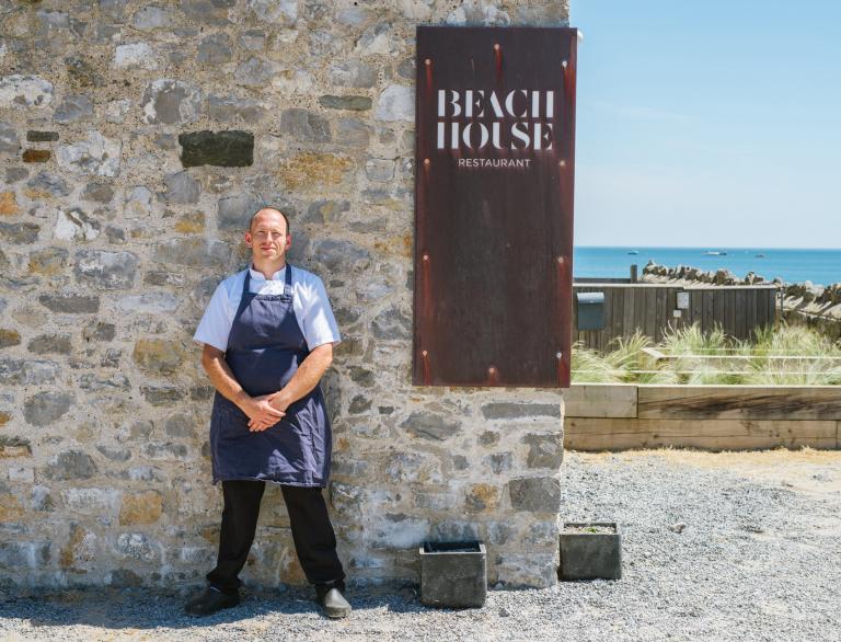 chef stood outside restaurant with beach in background.