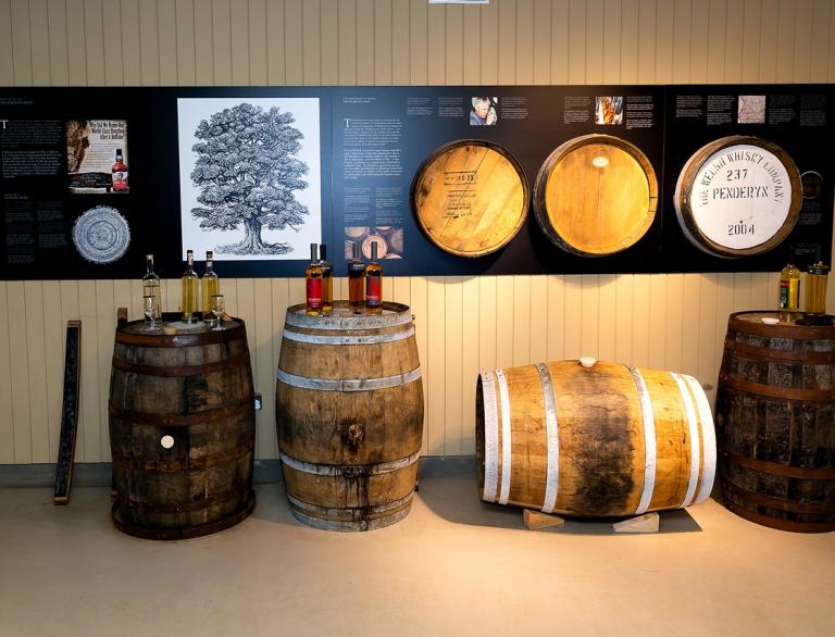 Whiskyfässer, auf denen Whiskyflaschen stehen, Informationstafeln hängen an der Wand dahinter.