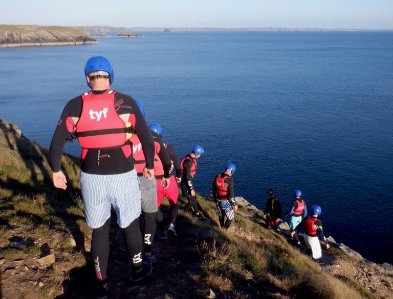 Coasteers wearing protective gear walking down a cliff toward the ocean.