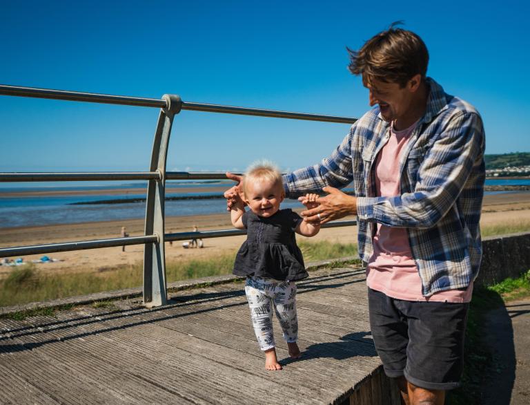 toddler walking on walkway held by man with beach in background.