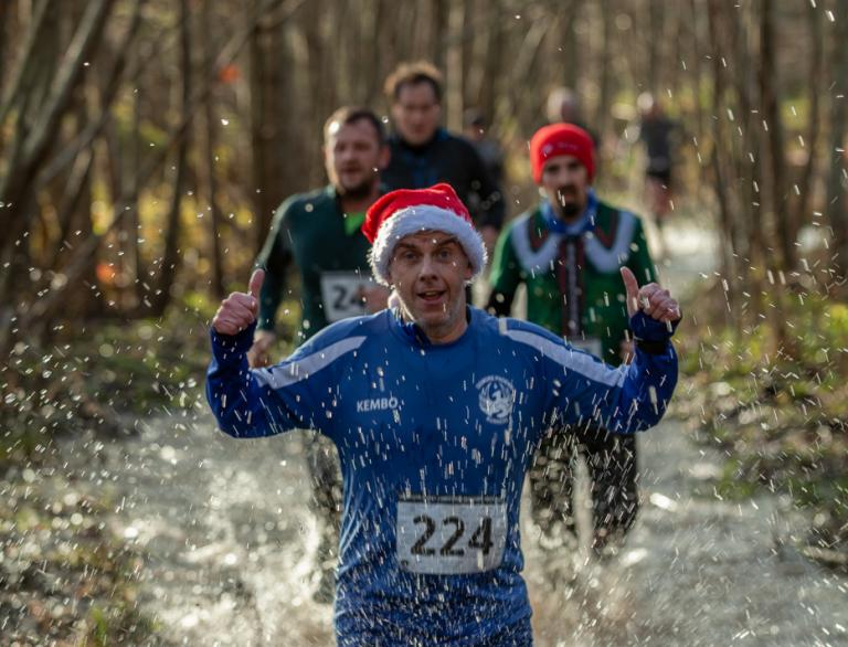 man with Christmas hat running through water, with other runners and bare trees in background.