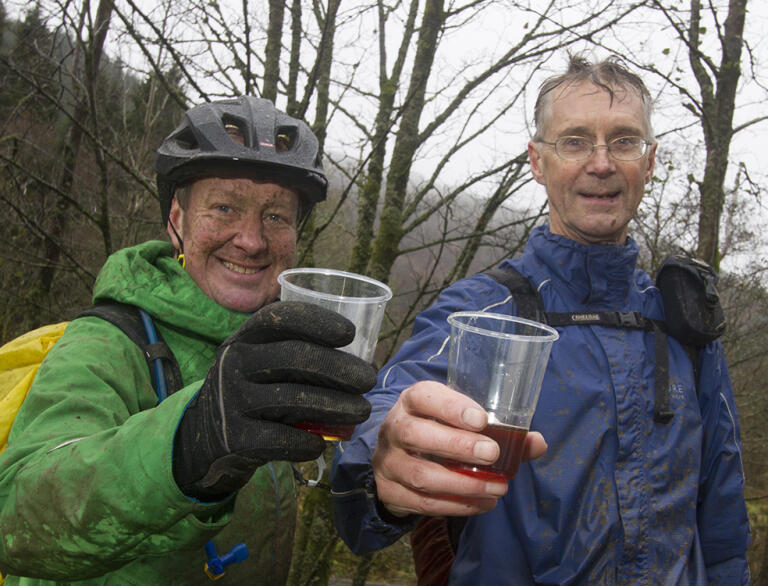 two men wearing muddy cycling gear and holding drinks.