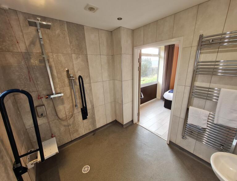 Barrierefreier Waschraum mit Dusche und Sitz sowie weit geöffneter Tür zum Schlafraum.