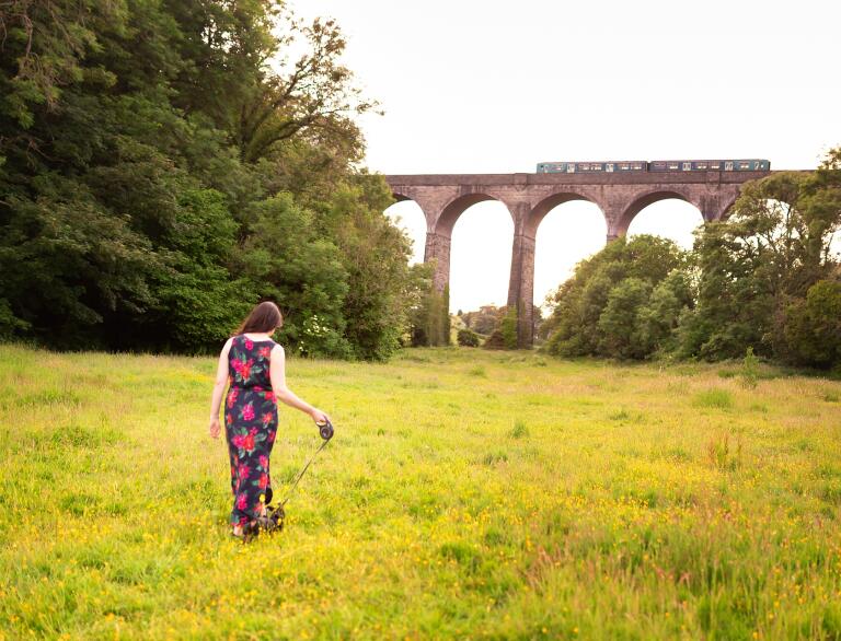 woman walking dog on grass, with viaduct in background.