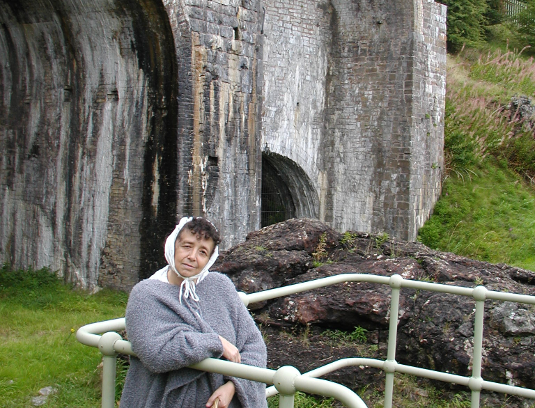 A woman in a headscarf and grey cloak in front of industrial works.