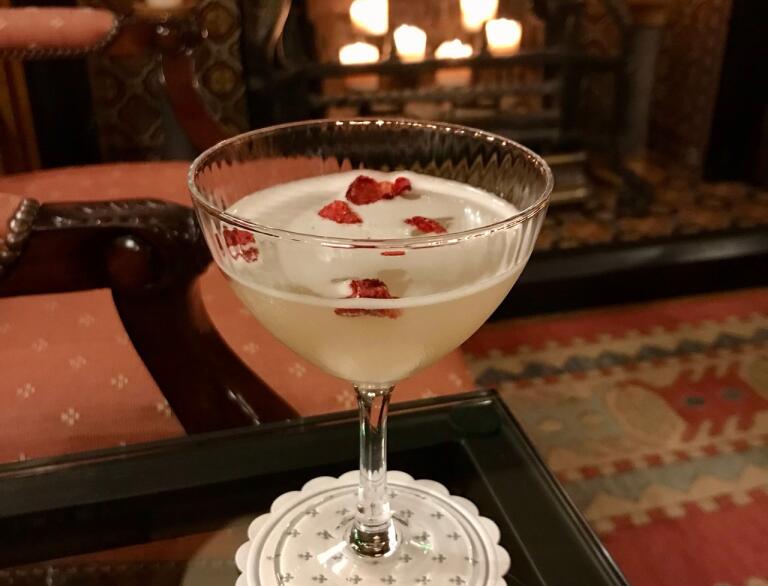 Ein heller Gin-Cocktail auf einem Beistelltisch, vor einem Kamin mit großen Kerzen.