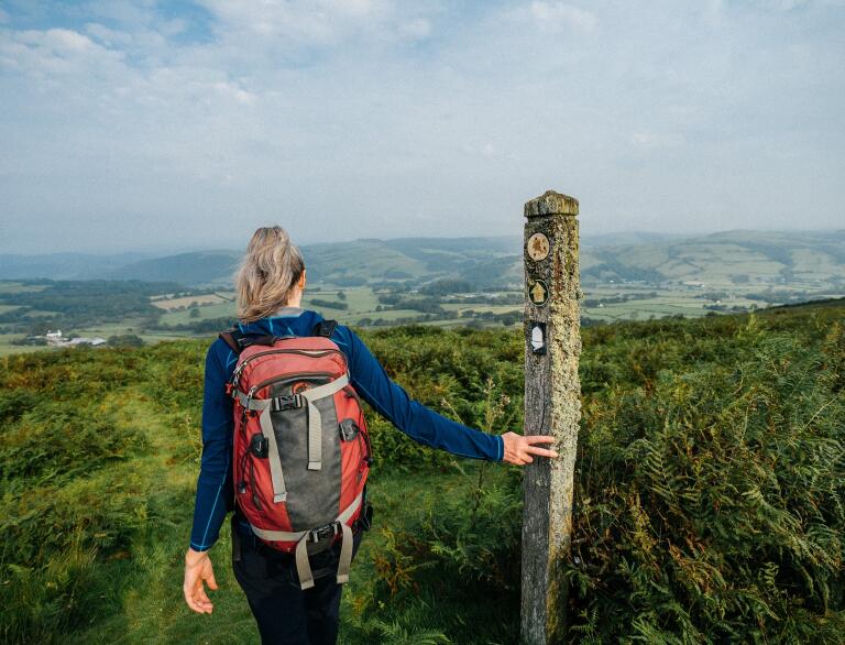 A female hiker on a path by a wooden waymarker, looking out over rolling green hills from above. 
