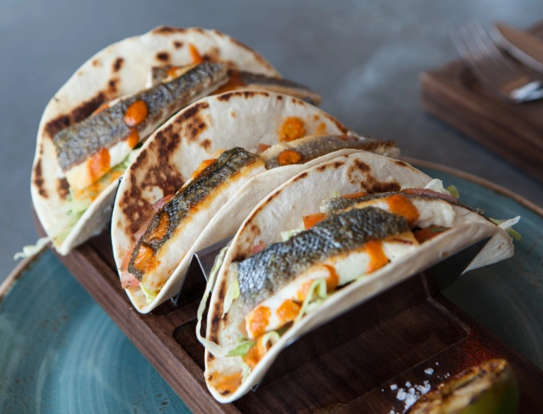 Three tacos filled with fish and lettuce and drizzled with an orange sauce. 