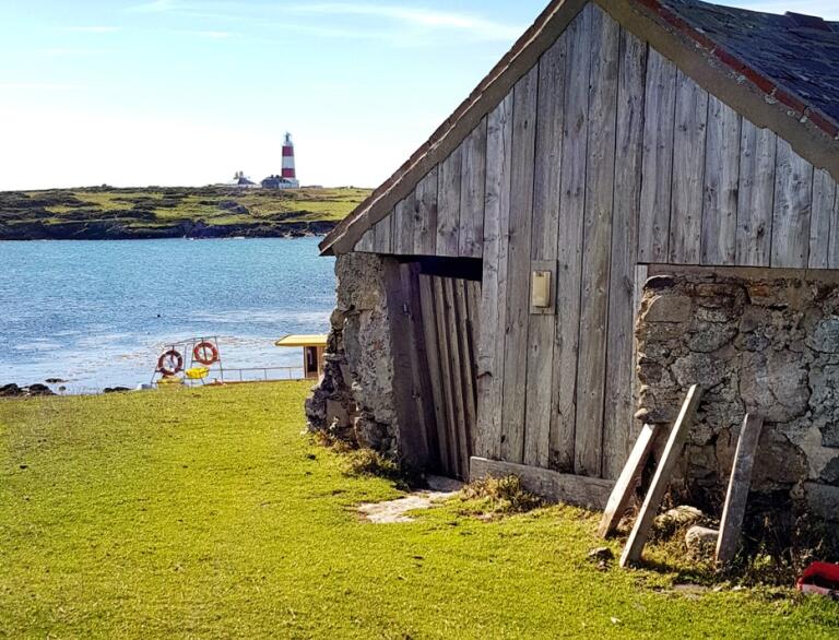 A wooden hut with green grass in the foreground and a lighthouse and water behind it.