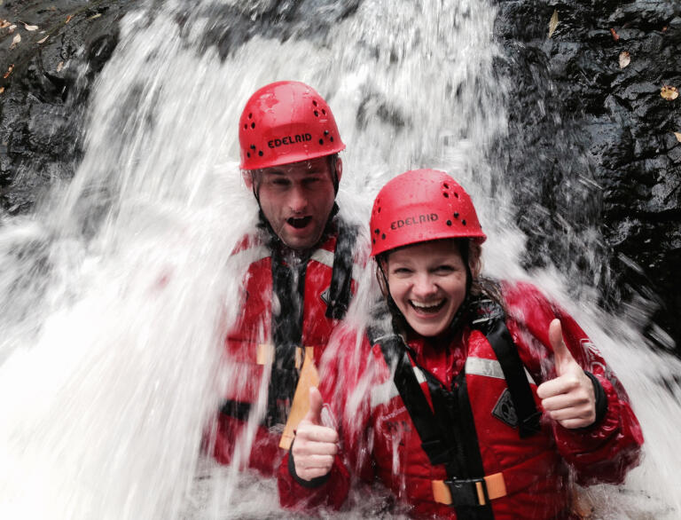 Two adults with red safety helmet and buoyancy aid standing under a waterfall. Female has thumbs up.