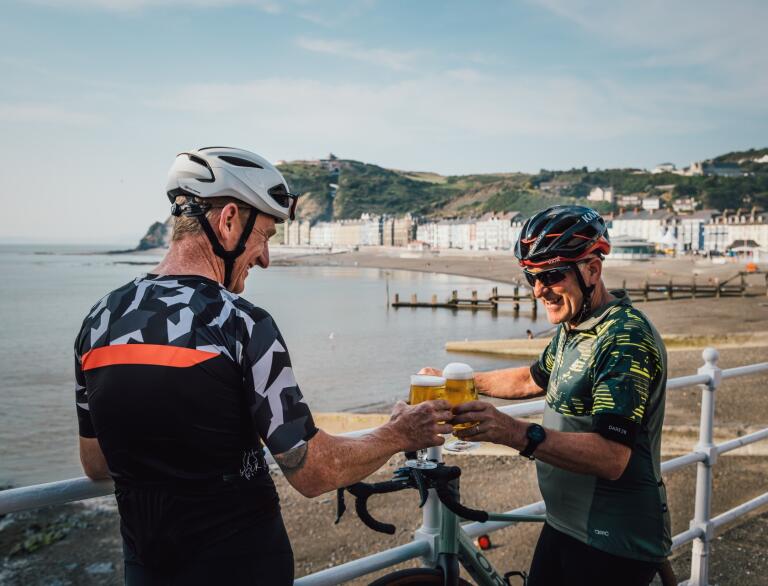 Two male cyclists raising a pint on a seaside prom with their bikes resting against railings.