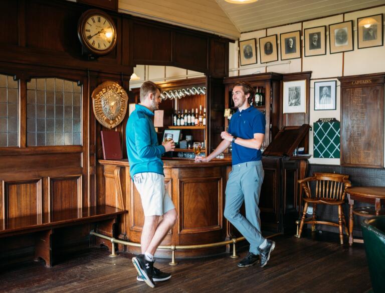 Two men standing by a bar in a dark panelled golf clubhouse.