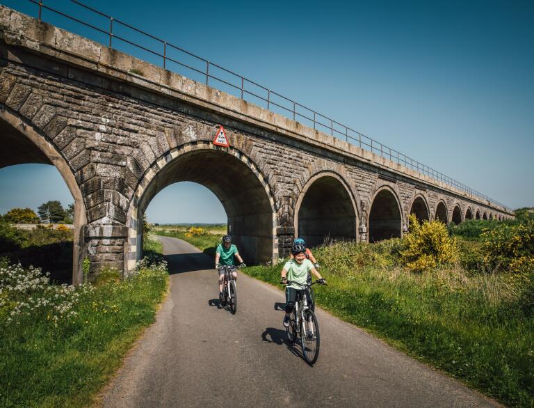 Two young cyclists on a flat path under a railway bridge. 