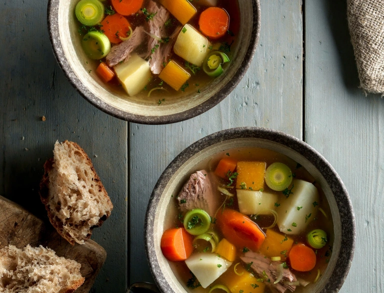 two bowls of cawl with bread and spoons on wooden table.