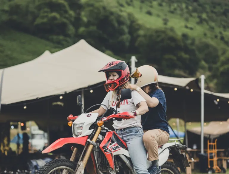 Two people on a motorbike in front of a festival tent in lush green mountains 