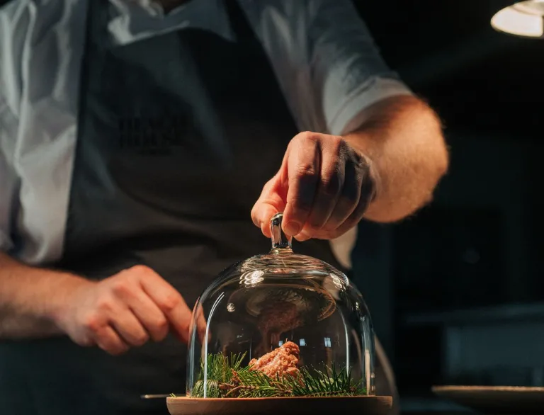 A male chef putting a small glass dome over a plate of food.