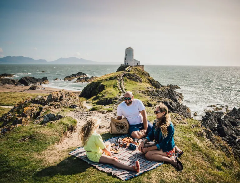 Two adults and a child having a picnic on a grassy cliffside, with a large white lighthouse in the background.