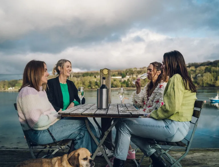 Four women sharing a bottle of wine on an outside table overlooking a beautiful strait.