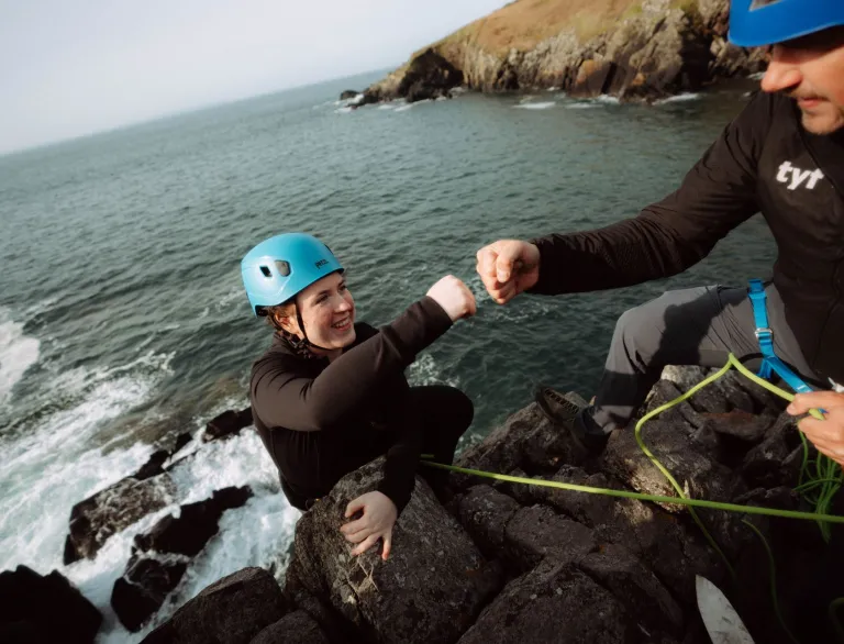 climber and instructor both wearing helmets fist bump on rock face with sea below.