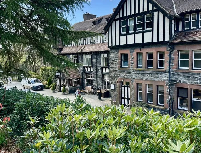Tudor-style multi-storey building with timber framing and white plaster, surrounded by lush greenery and trees under a partly cloudy sky