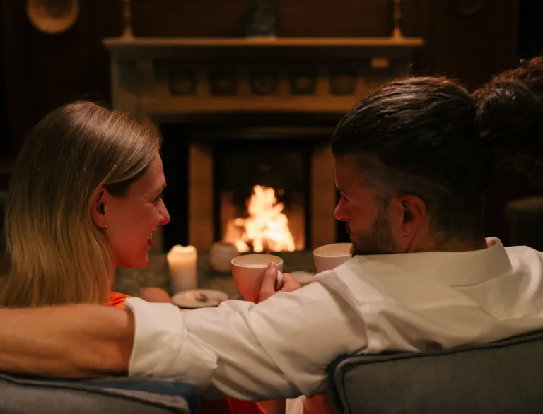 Couple seated on a couch by a lit fireplace, holding mugs and surrounded by candlelight in a cozy indoor setting.