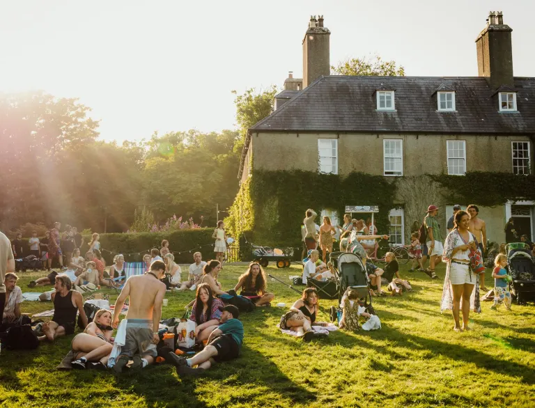crowd of people enjoying summer festival in garden with large house in background.