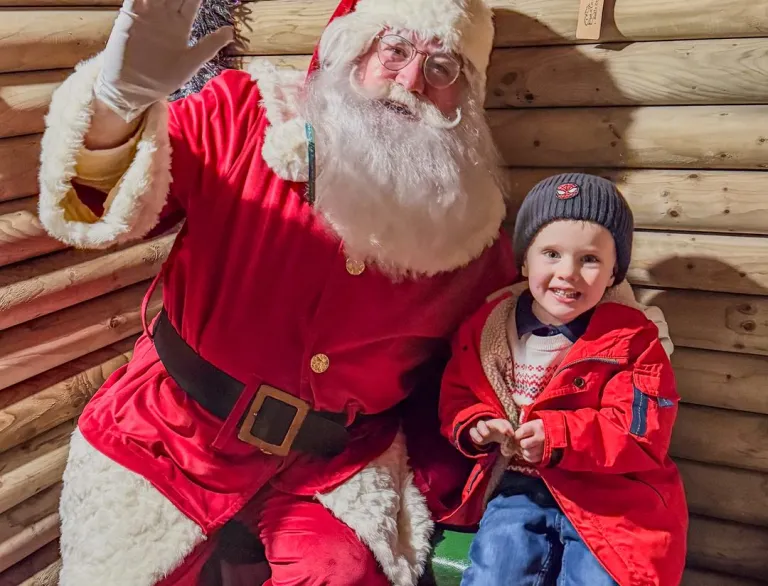 Father Christmas with hand in the air sat with a young boy.