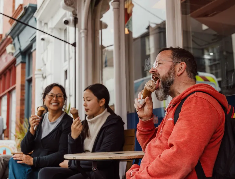 Three people sitting outside an ice cream shop on a high street, enjoying ice cream cones while seated at a small wooden table.