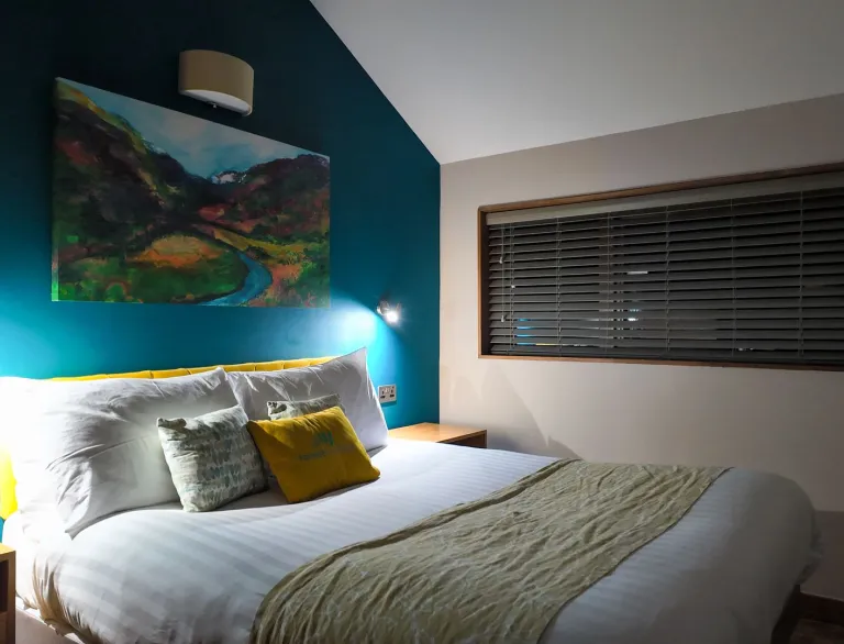 A modern bedroom with a double bed, teal feature wall and landscape painting above it.