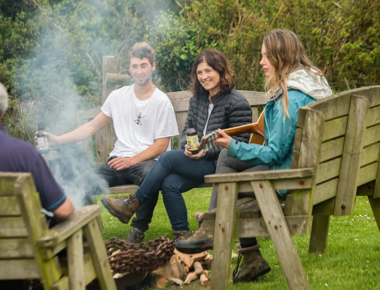 People sitting on wooden benches around a small outdoor fire pit, with smoke rising and greenery in the background; one person plays a guitar while others hold drinks.