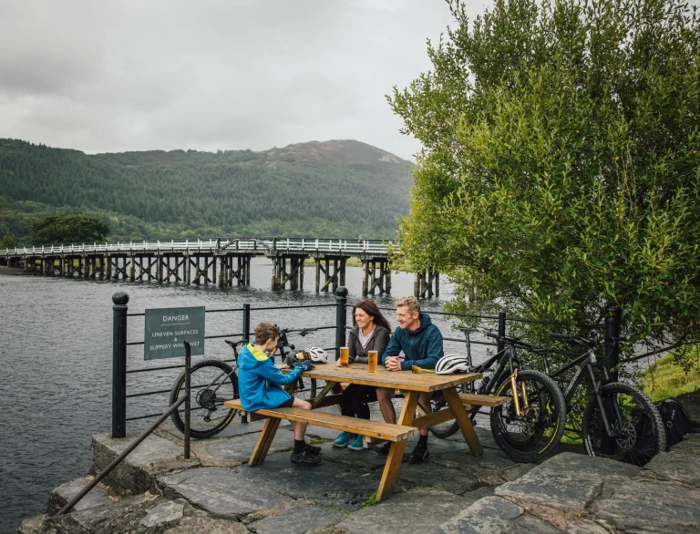 Two adults and a child sitting on an outside table, next to a wooden bridge over a river.