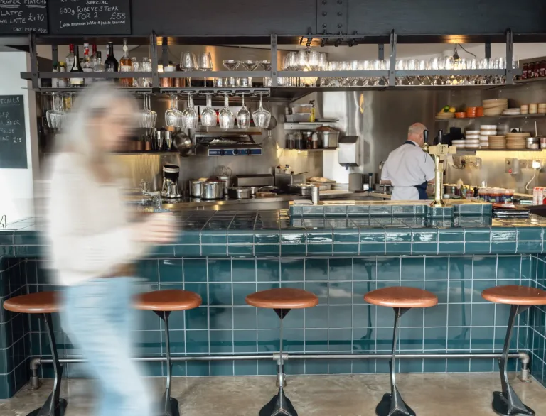 Interior of The Touring Cub in Penarth, featuring a teal-tiled bar counter, open kitchen, hanging glassware and bar stools, with a customer walking past in the foreground.