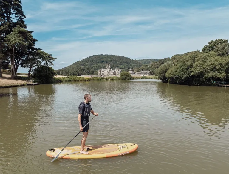 Person paddleboarding on a lake at Margam Park, with Margam Castle and wooded hills in the background beneath a blue sky.