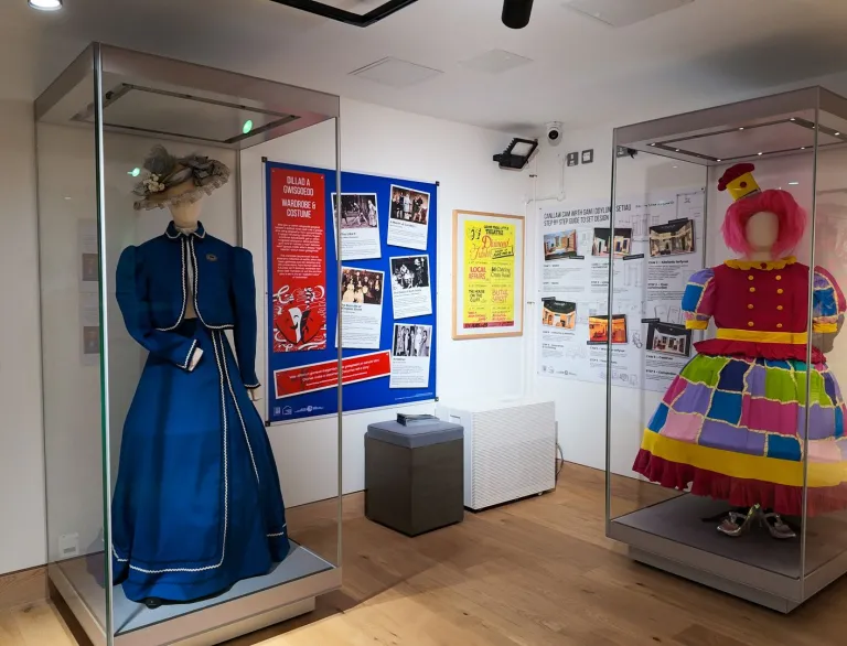 Two costumes displayed in glass cases at Wrexham Museum, including a blue Victorian-style dress and a colourful stage costume, as part of an exhibition on local theatre and performance history.