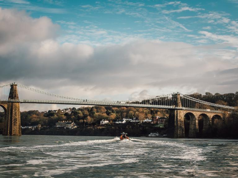 Image of the suspension bridge with houses and trees in the distance, and a rib boat on the Menai Strait