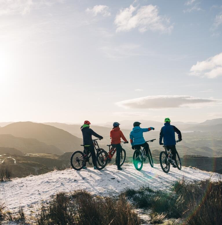 Vier Freunde auf Mountainbikes auf einer schneebedeckten Bergkuppe mit Blick über eine Berglandschaft.