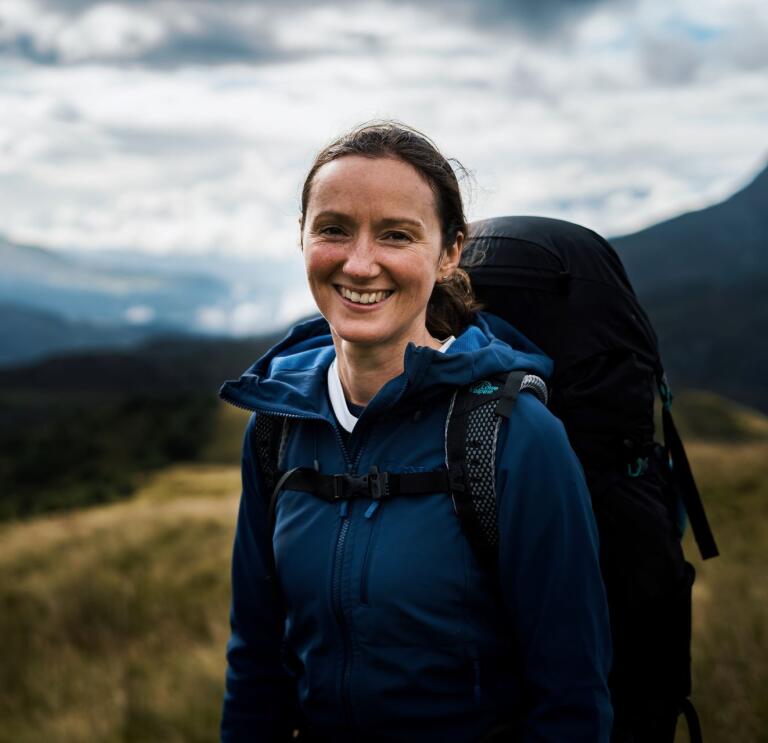 Claire Copeman smiling at the camera wearing outdoor gear and carrying a backpack with mountains in the background.