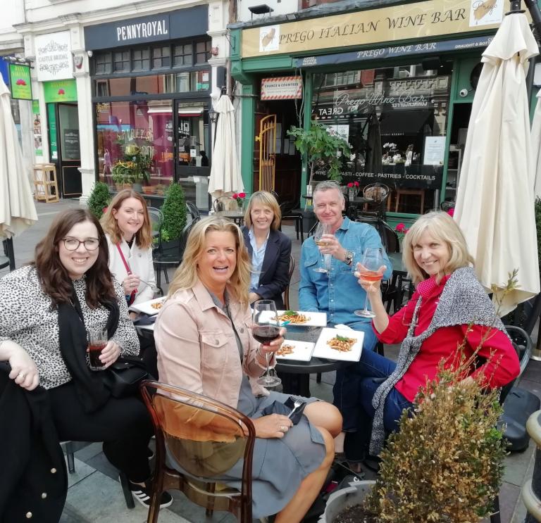 A group of people sitting outside an Italian restaurant raising a glass at the camera.