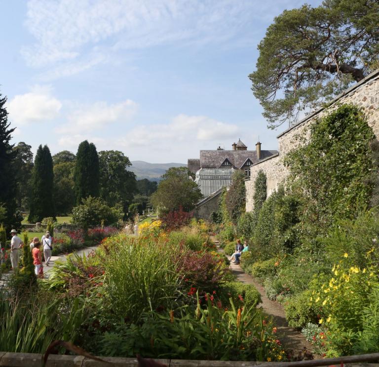 Flowers, trees and shrubs alongside paths and stone walls with a grand house in the background.