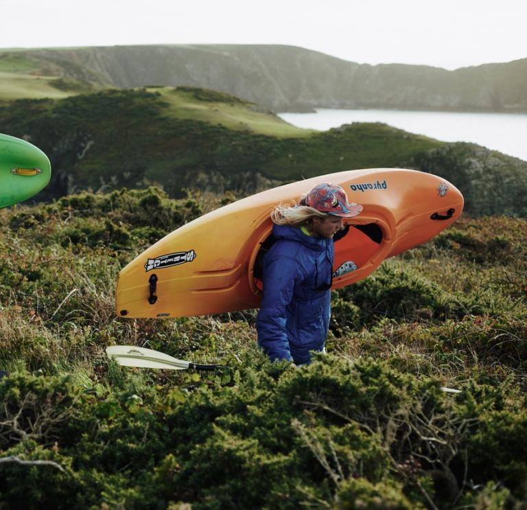 Two people walking through green foliage on a coastal path carrying a kayak each, heading to the sea.