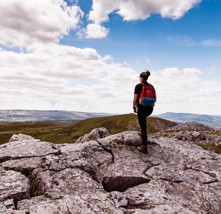 Wanderin auf einem Hügel stehend mit einem blauen Himmel im Hintergrund und mit Blick auf die grüne, bergige Landschaft der Brecon Beacons, Mittelwales