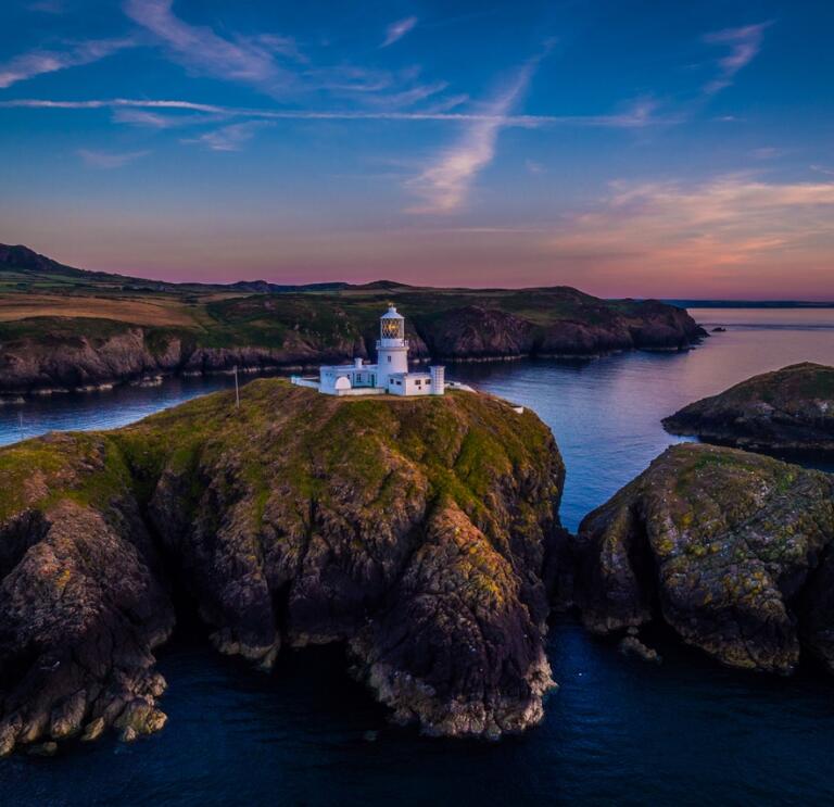 A lighthouse on a cliff with a a dramatic sunset behind.