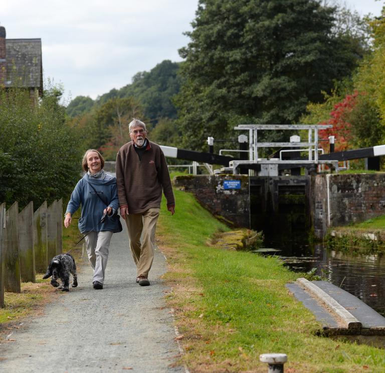 A woman, a man and a dog walking alongside a canal with a lock in the background.