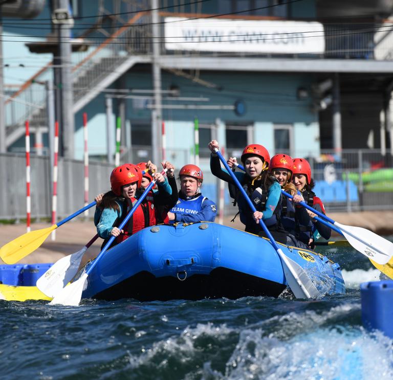 Eine mit Helmen und Schwimmwesten ausgerüstete Gruppe rudert in einem blauen Schlauchboot durch die Stromschnellen.